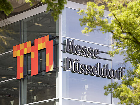 De buitenkant van een modern glazen gebouw met het logo ‘Messe Düsseldorf’ prominent op de gevel, gedeeltelijk omlijst door groene boomtakken.